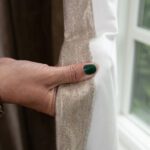 Hand with green nail polish and silver bracelet pulling back curtain beside window.