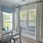 A sunlit dining room with a wooden table, chairs, and patterned curtains by a window.