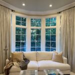 Elegant living room with large bay windows, beige drapes, and a white sofa.