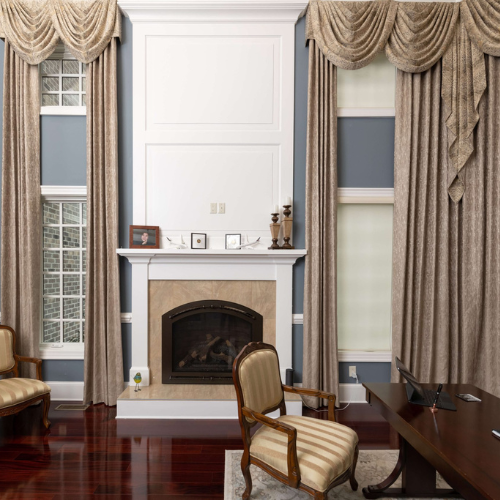 An office with a desk, fireplace and chair with grand curtains.